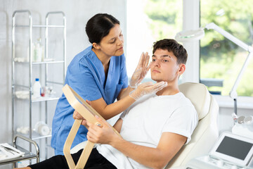 Young female dermatologist examining face of young male patient in clinic