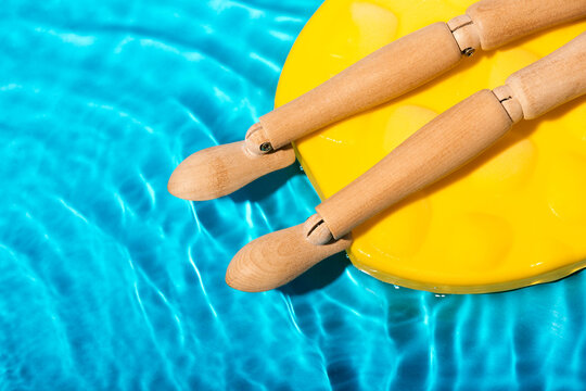 Body part of wooden mannequin figure sunbathing on a yellow pool float in clear blue swimming pool water