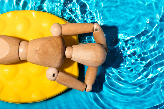 Wooden artist's mannequin relaxing on a yellow pool float in clear blue swimming pool water, simulating a summer leisure scene