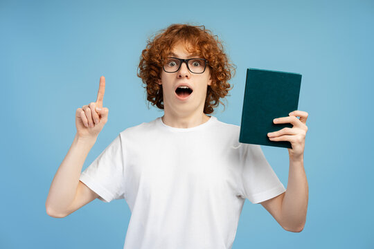 Excited teen in glasses, demonstrating a book and making index finder gesture on blue background