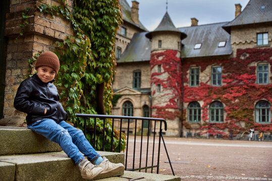 Kid smiling by a Swedish old university in a castle with red ivy climbing a brick wall in Autumn. Child sitting by the entrance of a Scandinavian botanical college conveys future campus life concept