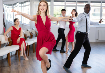 Positive adult woman in vibrant red dress enjoying active dances with black male partner in modern...