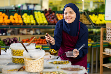 In grocery store asian female seller in hijab carefully puts large pickled olives in disposable...