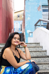 Portrait of beautiful young woman, with long black hair, sitting on a cement staircase against houses in the background.