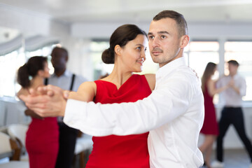 Concentrated elegant young adult man enjoying slow foxtrot with stylish female partner in red dress in dance studio. Amateur social dancing concept