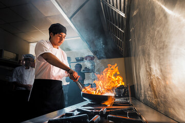 Young Asian chef in the restaurant kitchen making flambe in the frying pan. Cooking food in a frying pan with fire. Chefs working in the kitchen