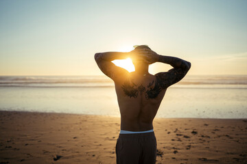 Chico joven musculado y tatuado posando en la playa sin camiseta al atardecer