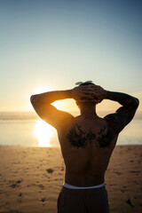 Chico joven musculado y tatuado posando en la playa sin camiseta al atardecer