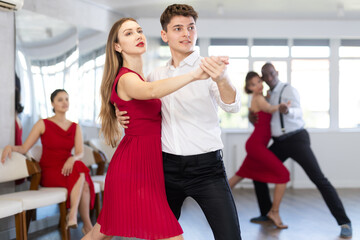 Positive modern young guy in formal attire and woman wearing elegant red dress gracefully waltzing...