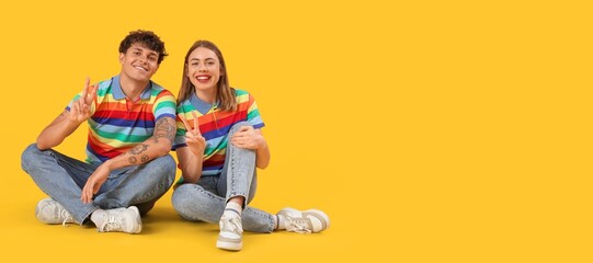 Young couple in rainbow t-shirts showing victory gesture on yellow background with space for text
