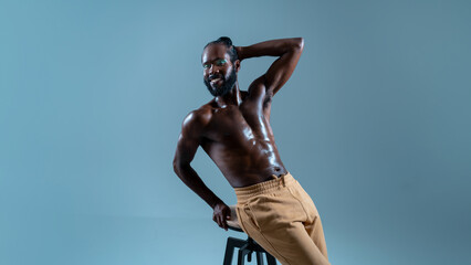 Fototapeta na wymiar Smiling shirtless gay man with bright makeup posing on stool. Happy muscular athletic African American man showing off his sexy body on blue background.