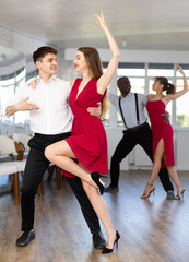 Fototapeta na wymiar Positive young Hispanic guy practicing passionate samba with stylish woman wearing red dress in dance class. Amateur dancing concept..