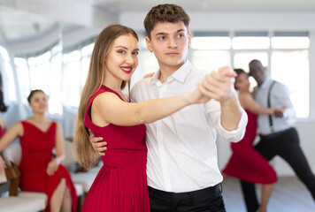 Pretty young woman dancing waltz with partner at group of multinational people in modern ballroom...