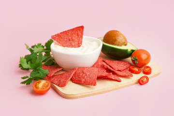 Wooden tray with red nachos, mayo and avocado on pink background