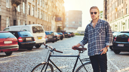 Young man holding his bicycle in the middle of a city street while wearing sunglasses
