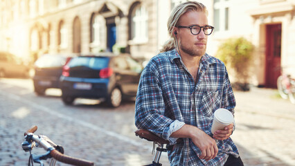 Young thoughtful man leaning on his bicycle on a street while holding a coffee to go