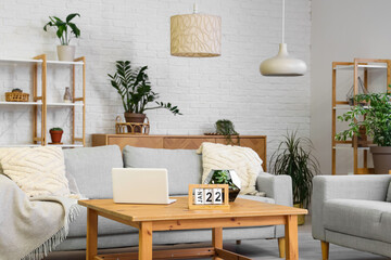 Stylish interior of beautiful living room with comfortable sofas, houseplants, shelving units and table with laptop