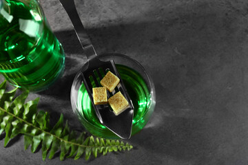 Absinthe in glass, spoon, brown sugar cubes, and fern leaf on gray textured table, above view with...