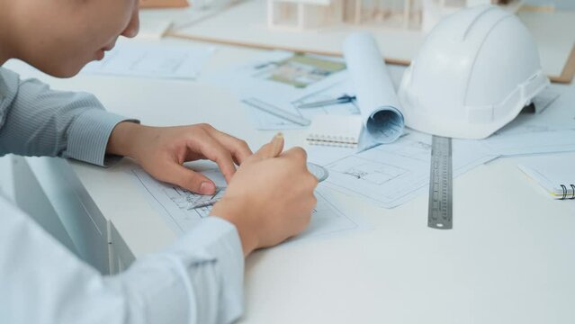 Close up of skilled architect engineer hand draws blueprint on meeting table with blueprint, architectural equipment, house model and safety helmet scatter around. Focus on hand. Immaculate.