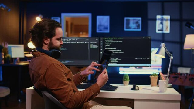 Software developer programming in home office, crosschecking lines of code between tablet and PC screen. IT admin holding portable device, using it as reference while coding on computer, camera B