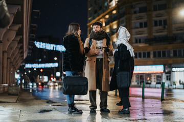 Three young adults stand in a lively discussion on a night-lit urban sidewalk, with city lights and traffic in the backdrop.