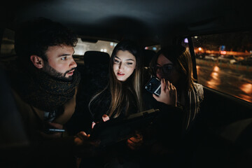 Three young adults are engaged in a collaborative effort, using a tablet and smart phone, while traveling by car in the city at night.