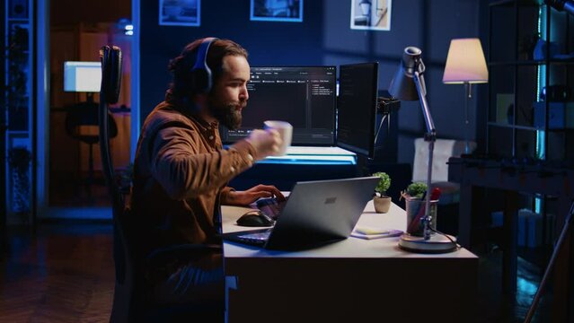 Programmer does multitasking to finish developing PC code, drinks coffee, listens music. IT admin wearing headphones, enjoying hot beverage, solving database errors on computer and laptop, camera B