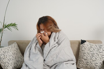 A sick Asian woman with flu sitting under the blanket. Her body was covered with a blanket lying on...