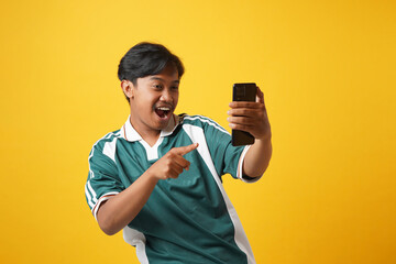 An Asian soccer fan emotionally watching a match football on a smartphone with a yellow background - obrazy, fototapety, plakaty