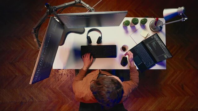 Top down view of engineer doing multitasking to finish developing PC code, drinking coffee, listening music. IT admin wearing headphones, solving database errors on computer and laptop, aerial shot