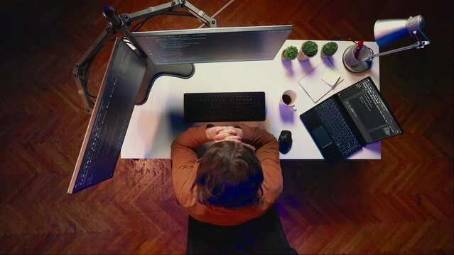 Aerial shot of computer scientist listening music while writing lines of code during remote job shift in home office. IT professional enjoying audio podcast, coding on desktop PC, top down view