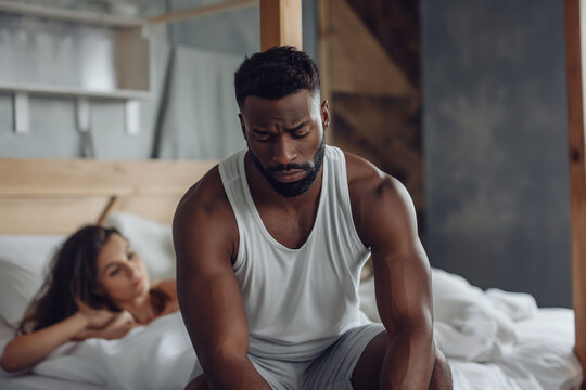 handsome black man sitting on the edge of his bed worried expression, in the background a woman looking at him. relationship problems