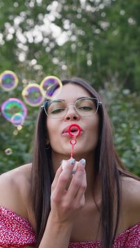 Portrait Happy Brunette Woman Wearing Glasses and in Pink Dress Blowing Soap Bubbles. Spring Time and World Women's Day. Vision Glasses Concept. Adult Plays Childhood. Vertical Video, Slow Motion.