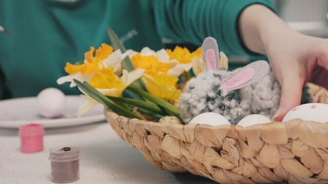 Close up of wicker basket with daffodils and handmade rabbit. Defocused woman decorating eggs for Easter on the background. The concept of preparing for the holiday