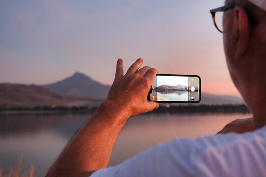 A man's hand holding a mobile phone captures the natural beauty of the sunset over the mountain.