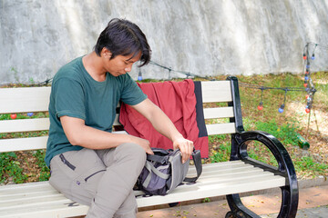 A handsome man is rummaging through his sling bag and sitting on a park bench.