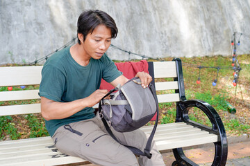 A handsome man is rummaging through his sling bag and sitting on a park bench.