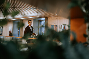 Riga, Latvia - January 20, 2024 - A groom in a modern setting, framed by foliage, holds a bouquet, with a stylish interior background.