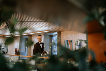Riga, Latvia - January 20, 2024 - A groom holding a bouquet, viewed through foliage, in a modern room with wooden walls and abstract art.