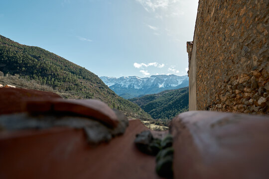 Snowy mountains in the background seen through the out-of-focus brown tiles of a roof with a stone wall on one side and a green mountain on the other