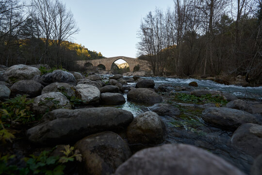 Old stone bridge in the distance with the foreground of stones next to a river surrounded by trees at sunset without clouds in the sky