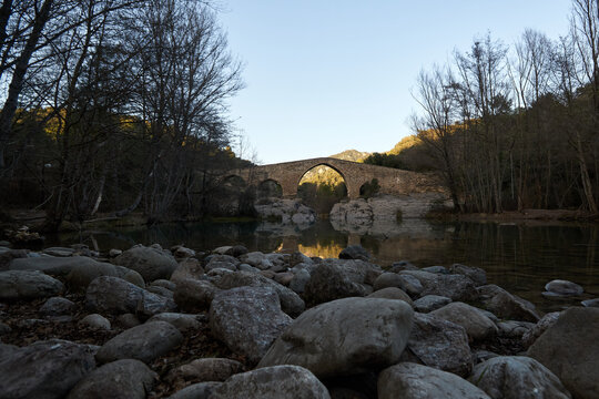 Old stone bridge in the distance with the foreground of stones next to a river surrounded by trees at sunset without clouds in the sky