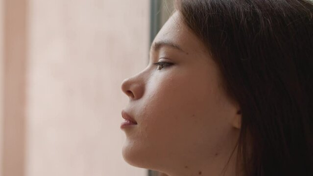 Woman looks through window sadly from indoors. Attractive lady handles with psychological issues alone at home. Frustrated woman overthinks situation