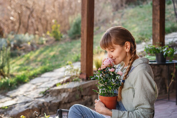 Young happy woman inhaling the scent of a rose in a pot while sitting on the terrace on a spring day - 743607046
