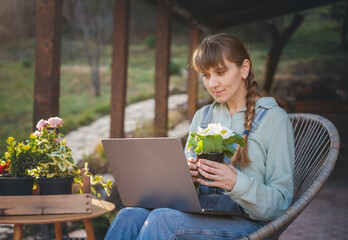 A young woman sitting on the terrace in front of a laptop with a flower in a pot in her hands, online gardening courses. Online floriculture training - 743608028