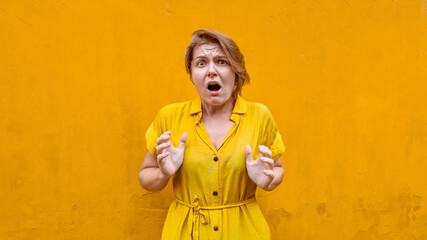 Shocked woman in yellow dress gesturing with her hands against a vibrant yellow background with...