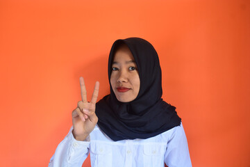 Asian Muslim woman close up raising two fingers with smiling face on blurred orange color textured background with copy space