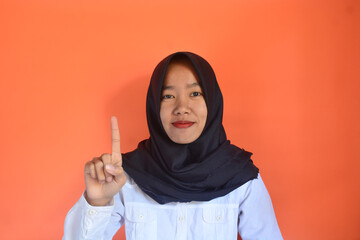 Asian Muslim woman close up raising one finger with smiling face on blurred orange color textured...