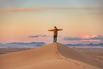 Happy young hiker is standing with open arms at sand dune and enjoys sunset sky