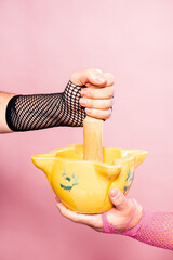 Close-Up of Two Hands Grinding Herbs in a Yellow Mortar and Pestle Against a Pink Background